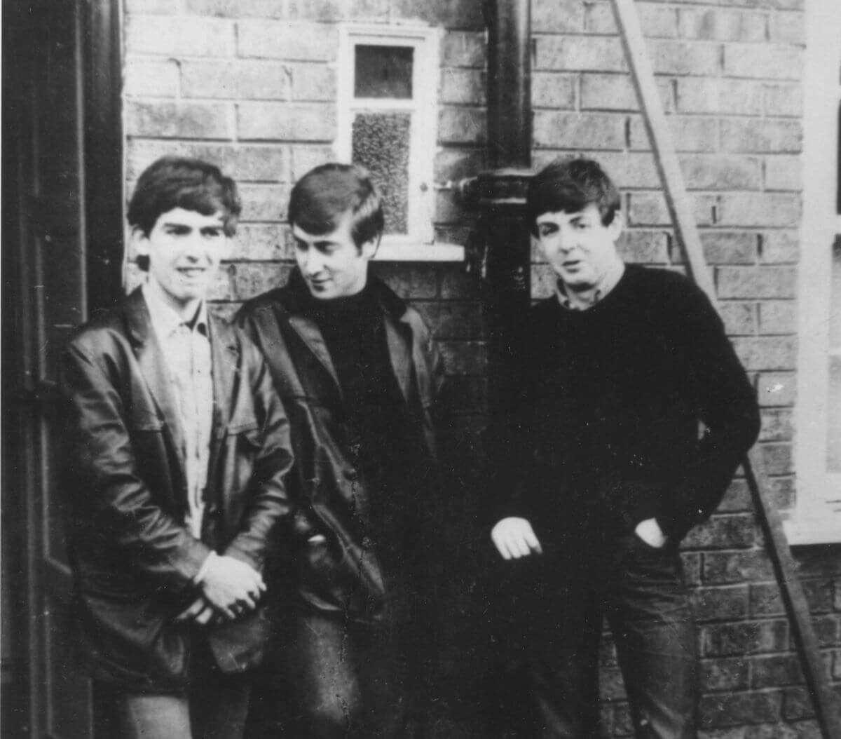 A black and white picture of George Harrison, John Lennon, and Paul McCartney leaning against a brick wall.