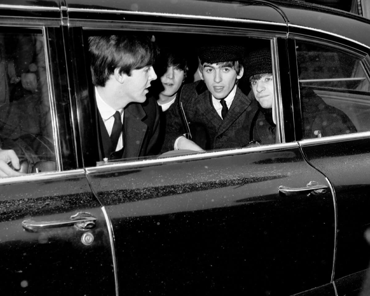 The Beatles wear suits and gather in the backseat of a car.