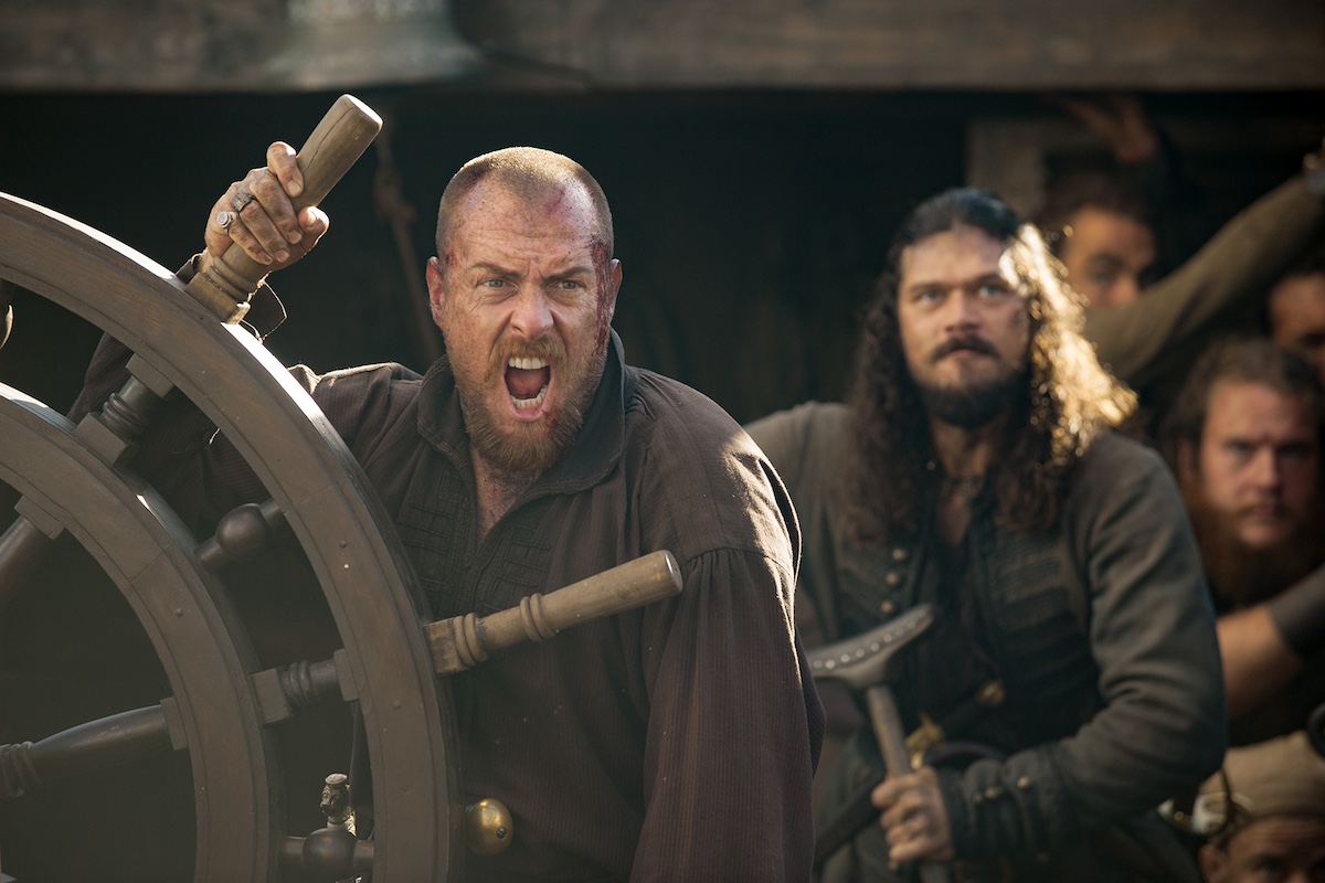 Toby Stephen with his hand on a ship's wheel next to a an with long dark hair and a beard in 'Black Sails'