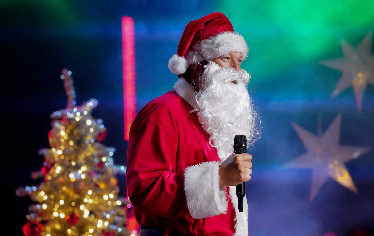 Blake Shelton dressed as Santa Claus holding a microphone with a Christmas tree in the background for a holiday episode of 'Barmageddon' Season 2