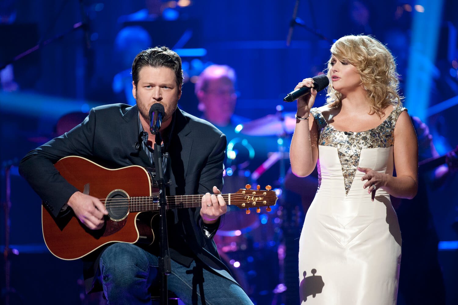 Blake Shelton playing guitar next to Miranda Lambert singing into a microphone on stage. Lambert wears a white sparkling dress.
