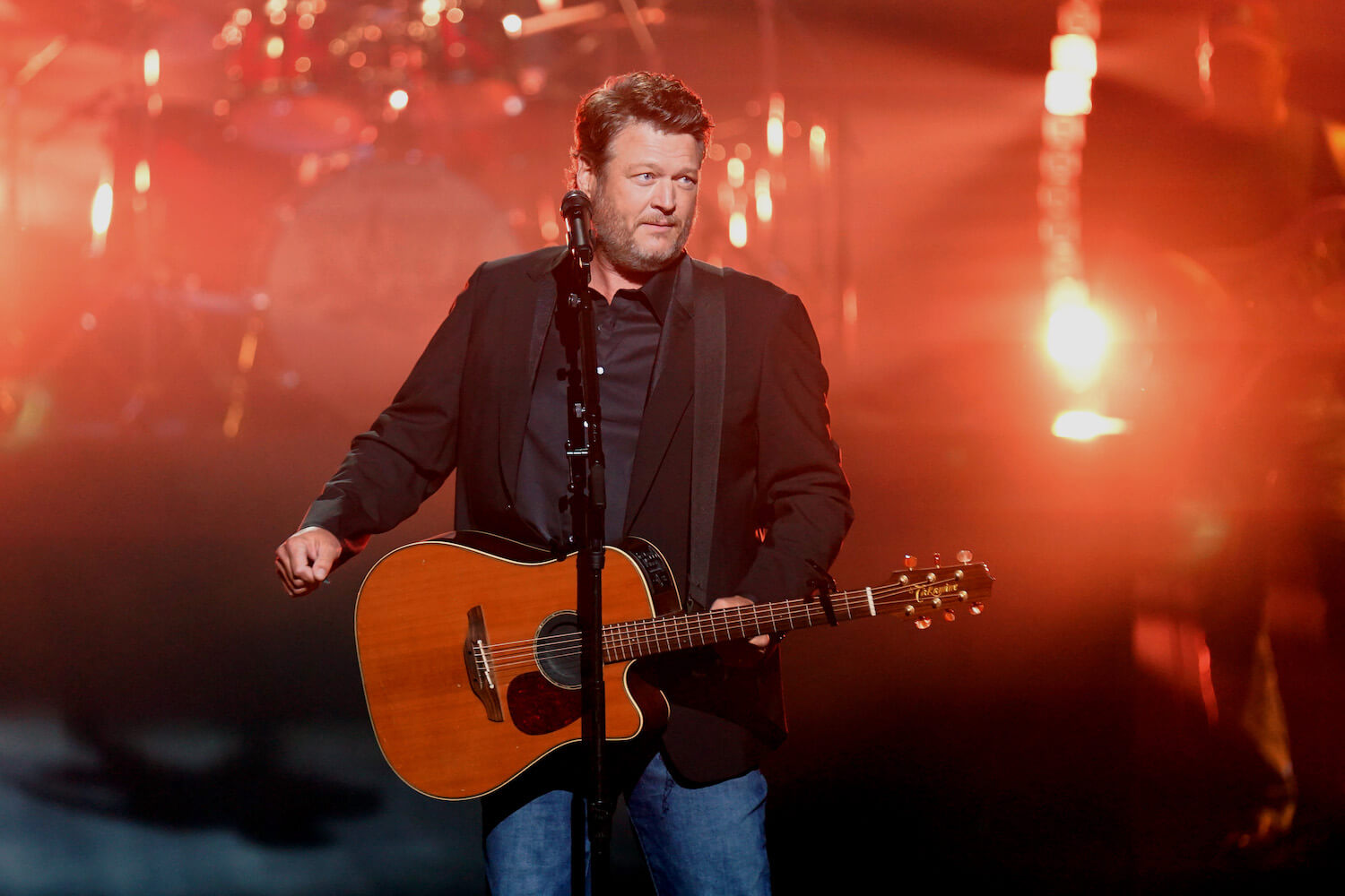 Blake Shelton with a guitar strapped around his neck on stage in front of orange lighting