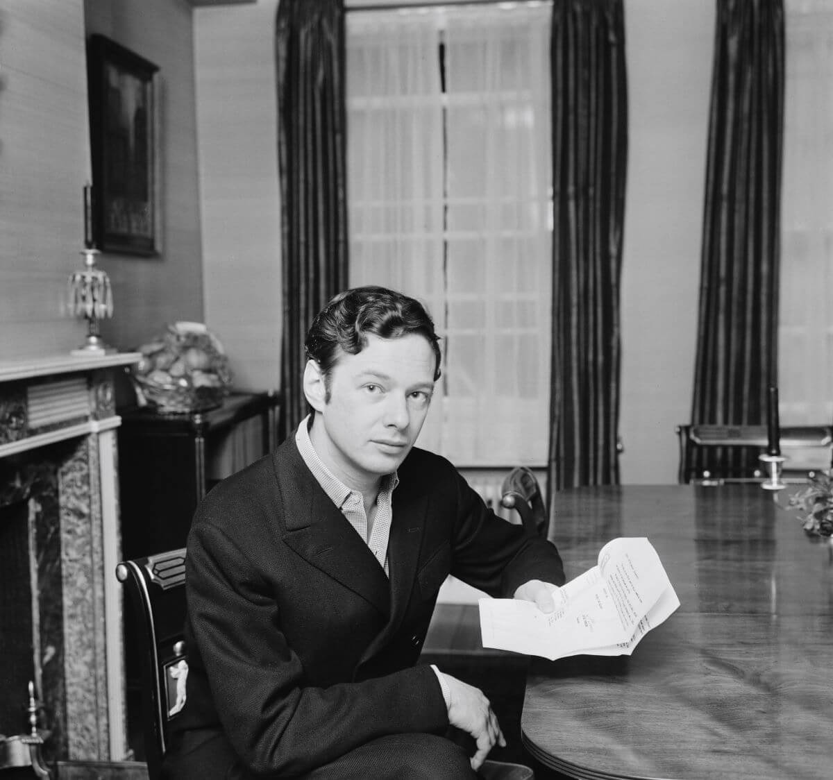 A black and white picture of Brian Epstein sitting at a table holding papers.