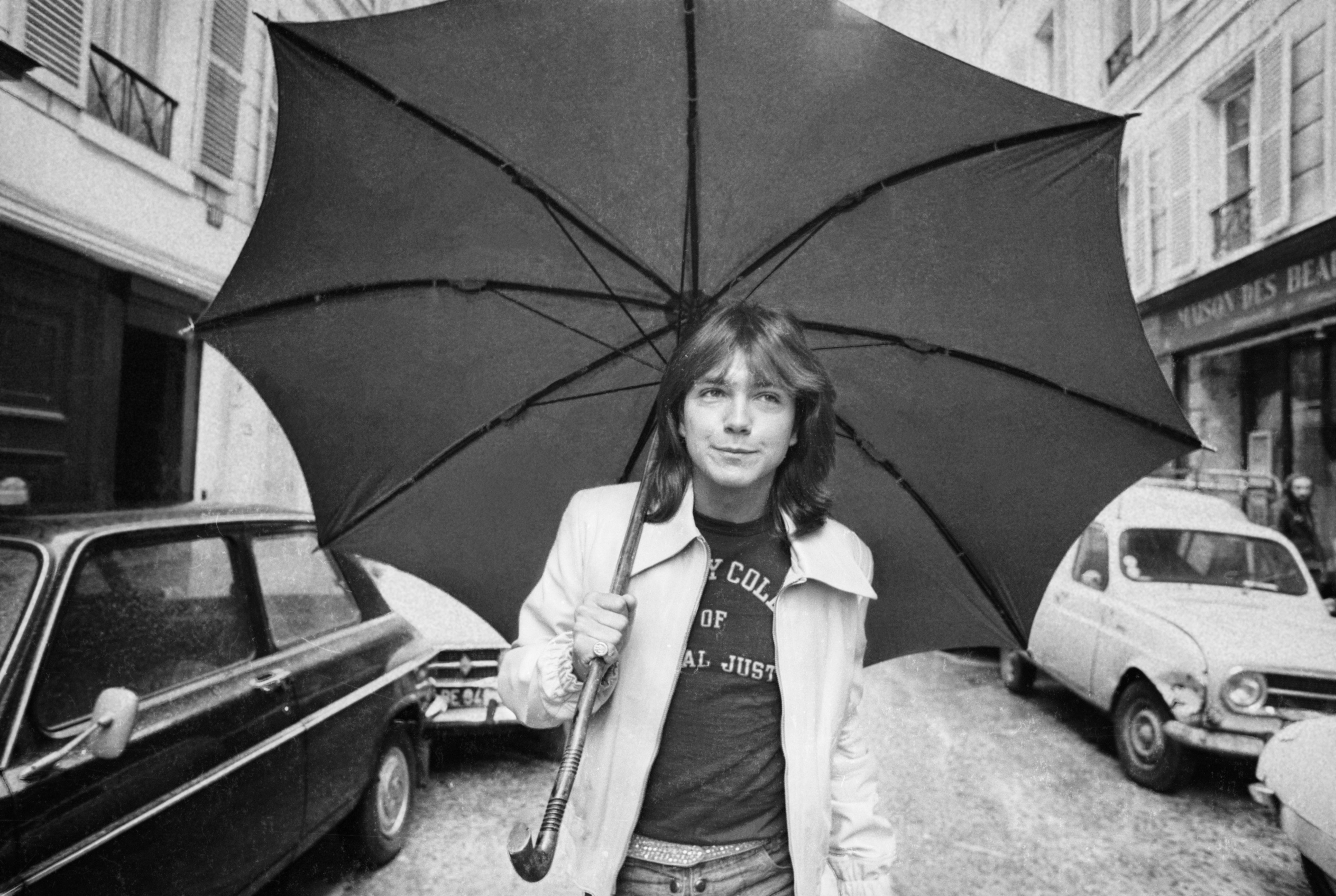 Black and white photo of David Cassidy of 'The Partridge Family' walking down a Paris street under an umbrella