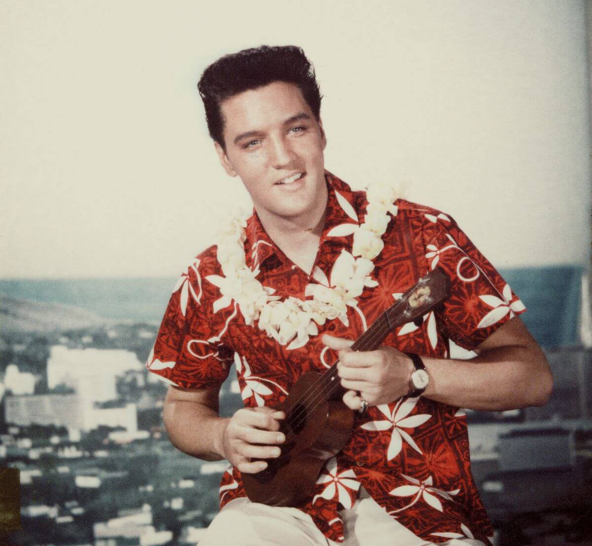 Elvis Presley wears a red shirt and a lei on the set of 'Blue Hawaii.' He strums a ukulele.