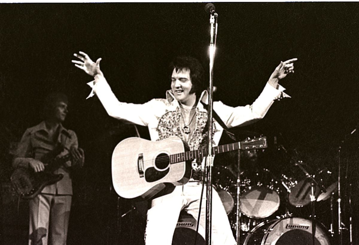 A black and white picture of Elvis Presley wearing a jumpsuit. He holds his arms up and has an acoustic guitar strapped to him. He stands behind a microphone. 