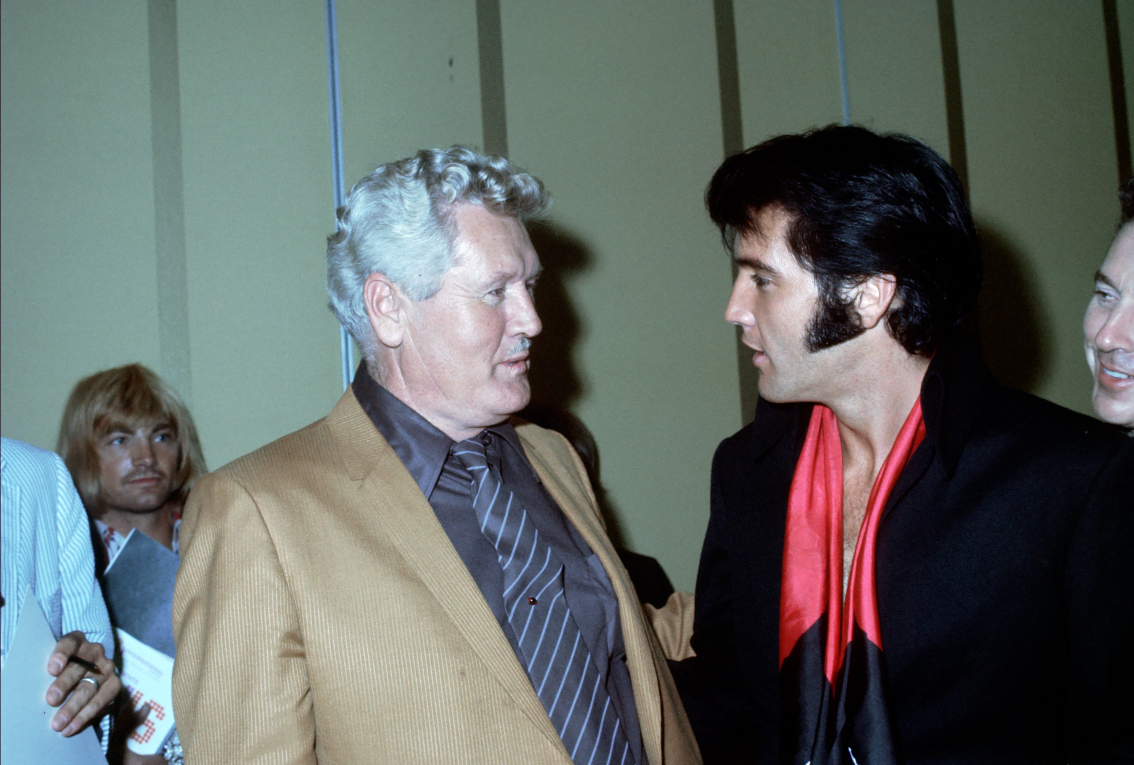 Vernon Presley wears a brown jacket and talks to Elvis Presley, who wears a red shirt and black jacket. There are several other people in the room.