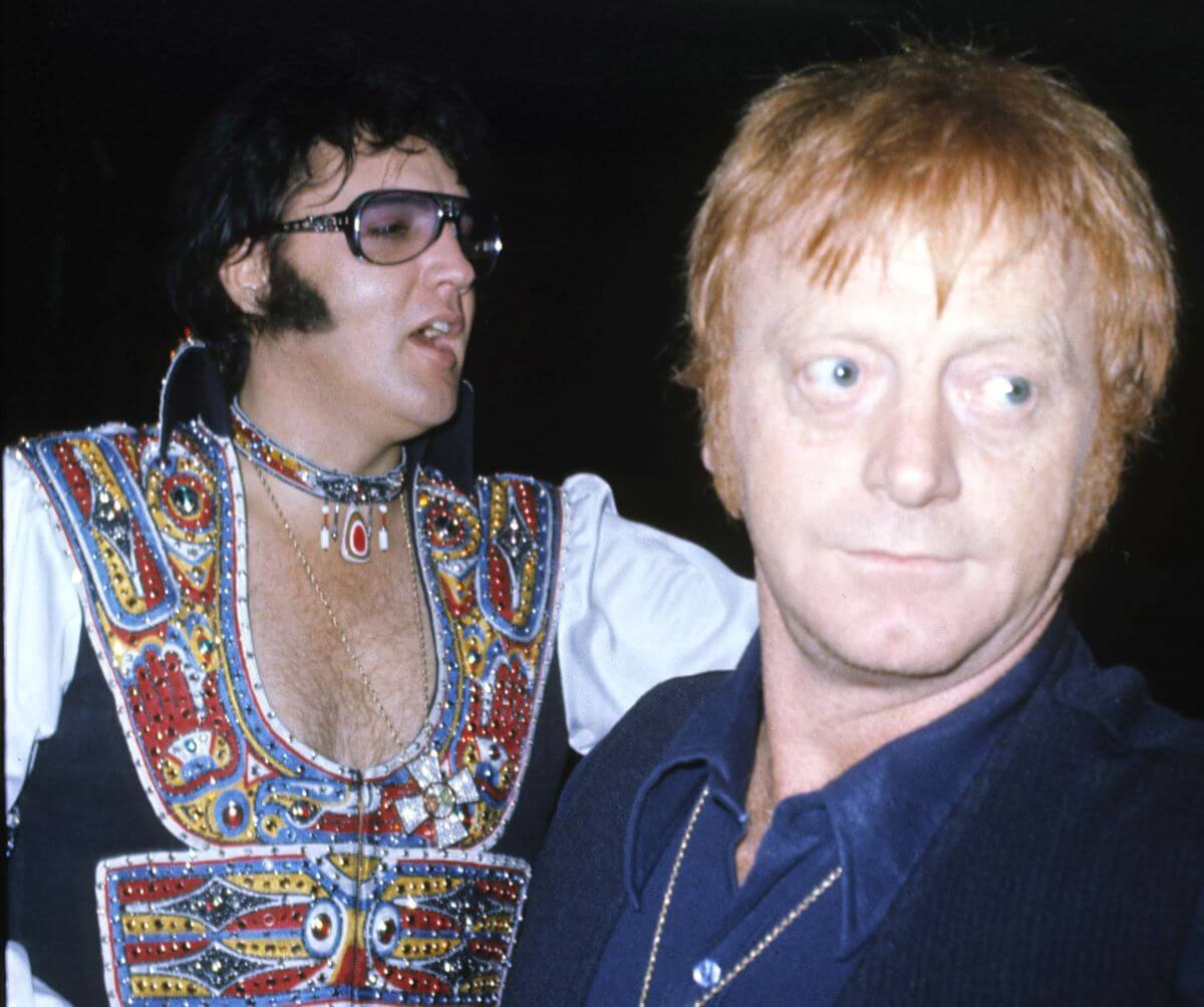 Elvis Presley wears sunglasses and a patterned shirt. His bodyguard, Red West, stands in front of him.