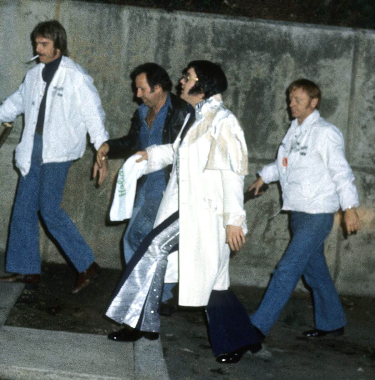 Elvis Presley walks in front of a concrete wall with three bodyguards. He wears a long white coat.