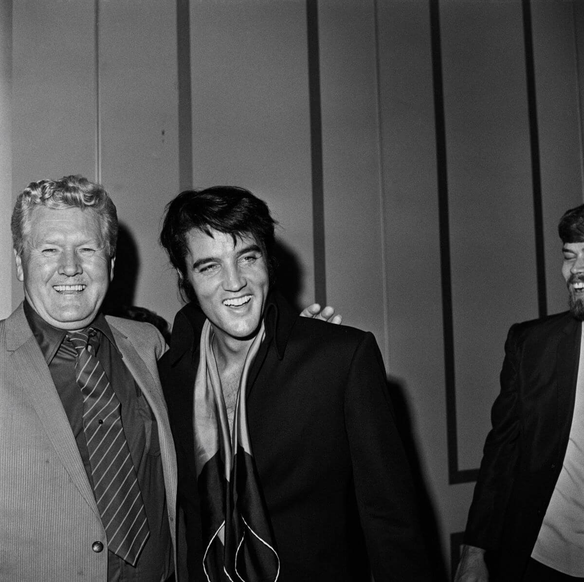 A black and white picture of Vernon Presley standing with his arm around Elvis' shoulders. They both smile.