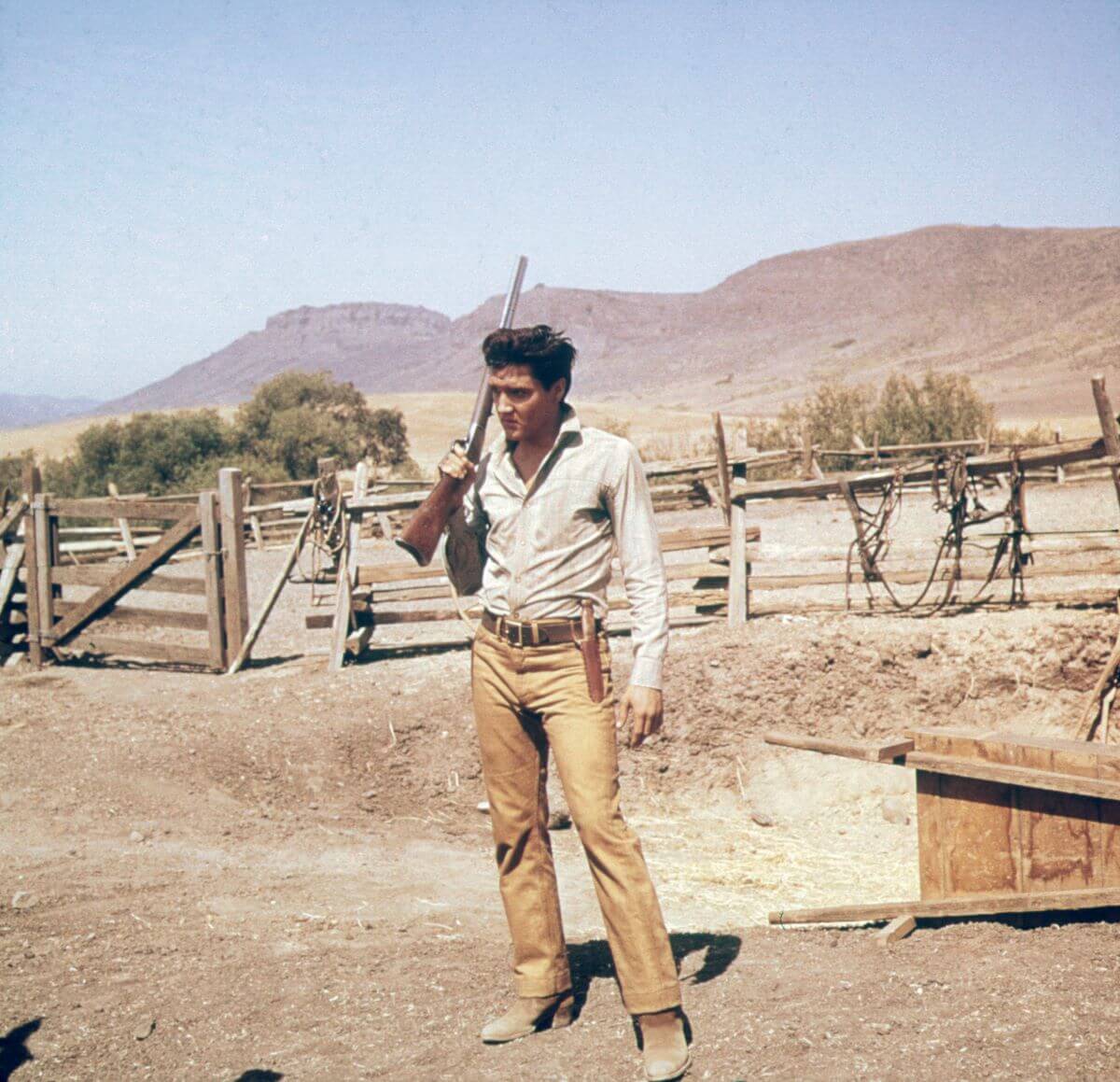 Elvis Presley holds a gun over his shoulder and stands outside. There are mountains and a fence behind him.