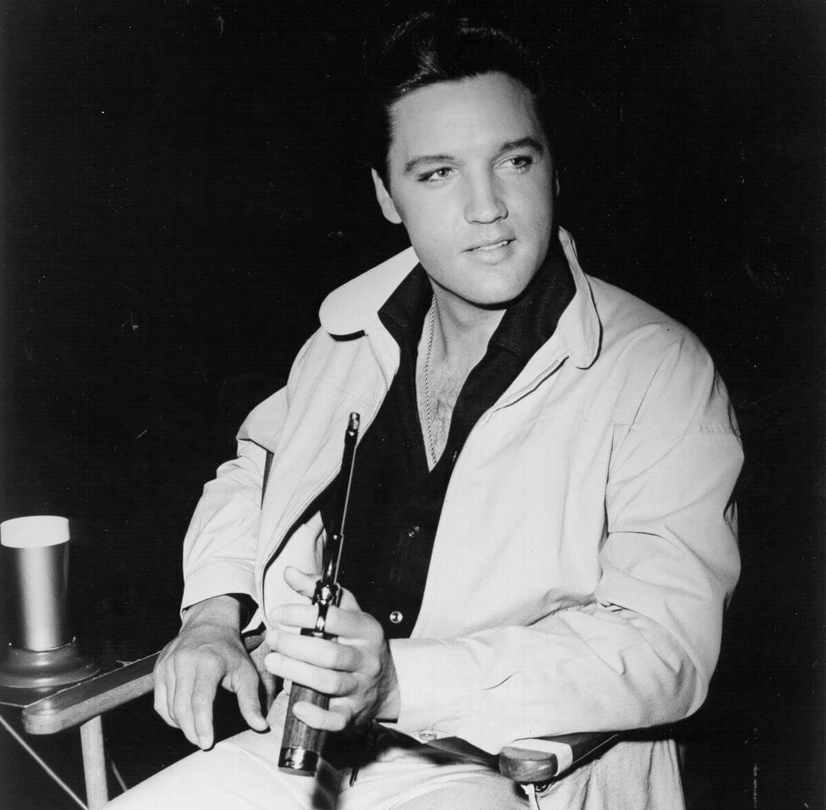 A black and white picture of Elvis Presley sitting in a chair and holding a gun in one hand.