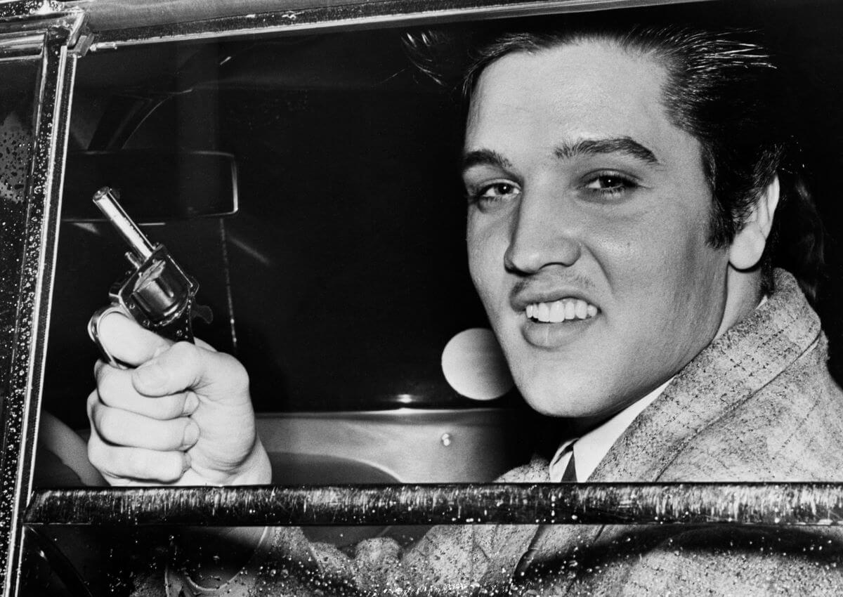 A black and white picture of Elvis Presley holding a gun. He sits in a car with the window rolled down.