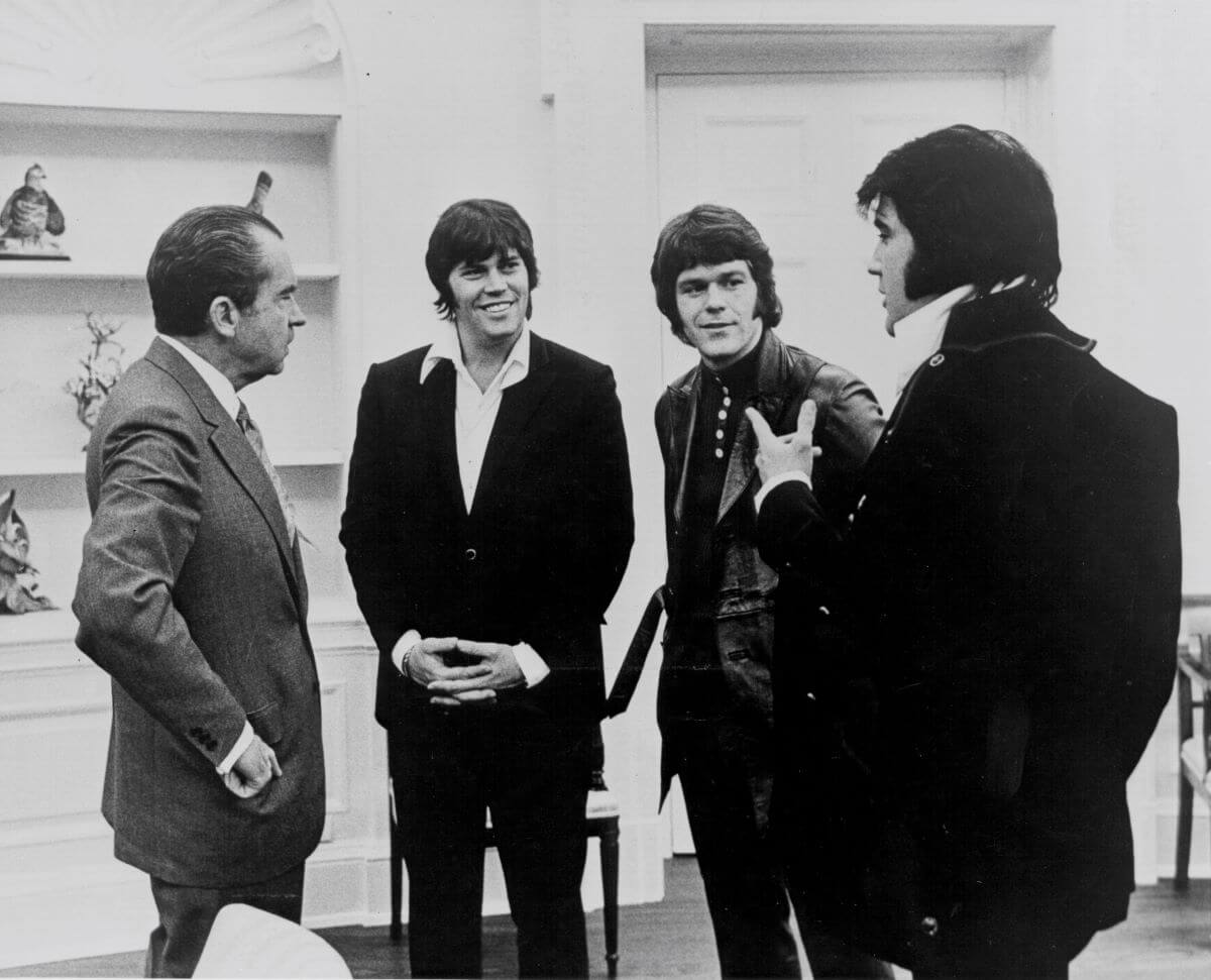 A black and white picture of Elvis Presley in the Oval Office  with Richard Nixon, Sonny West, and Jerry Schilling. Elvis' back is mostly to the camera.