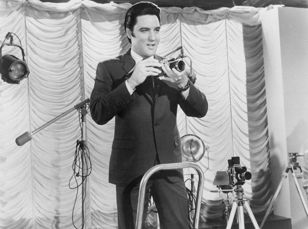Elvis Presley stands in front of a curtain and film equipment and holds a camera.