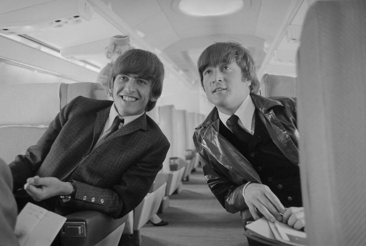 George Harrison and John Lennon lean toward each other across the aisle of an airplane.