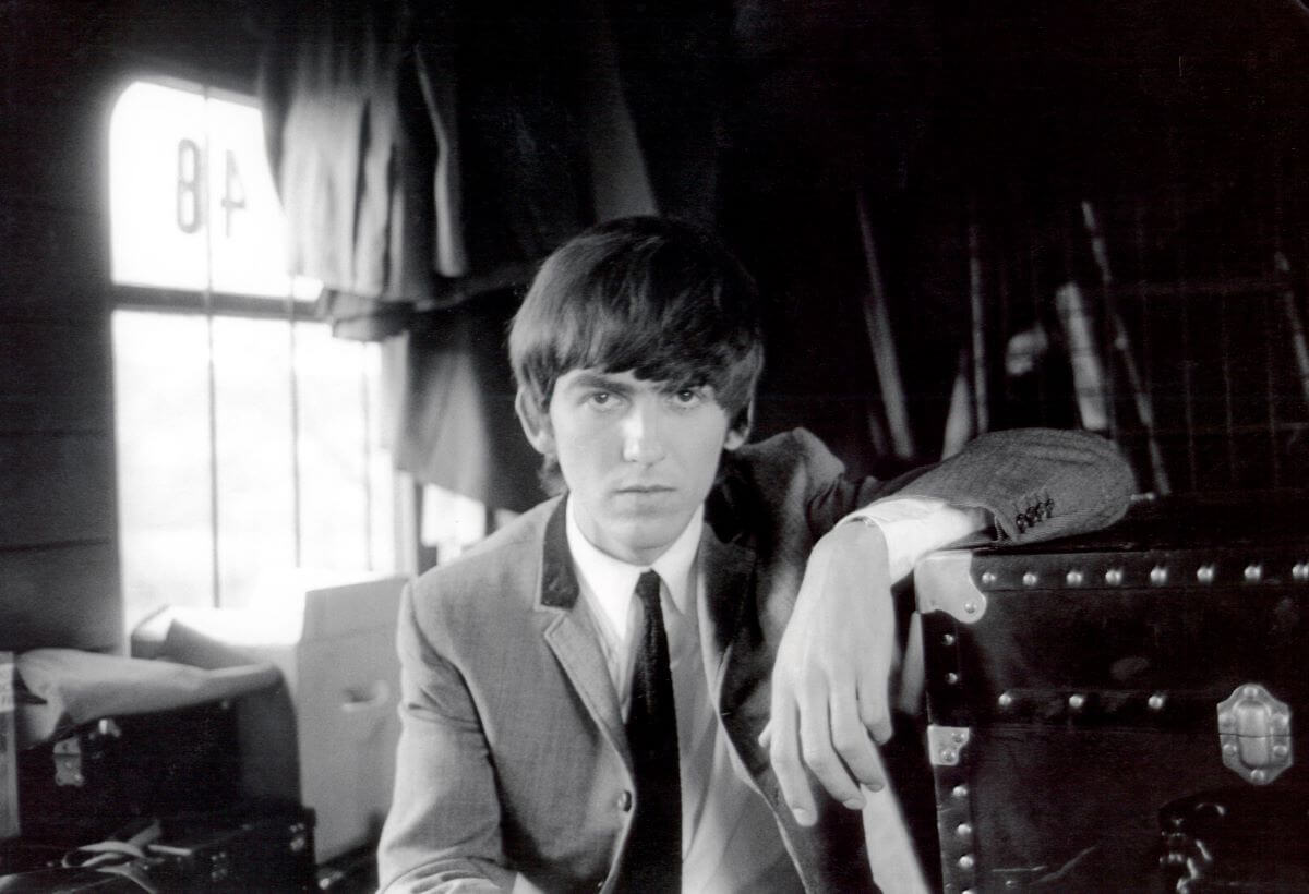 A black and white picture of George Harrison wearing a suit and resting his arm on a trunk.