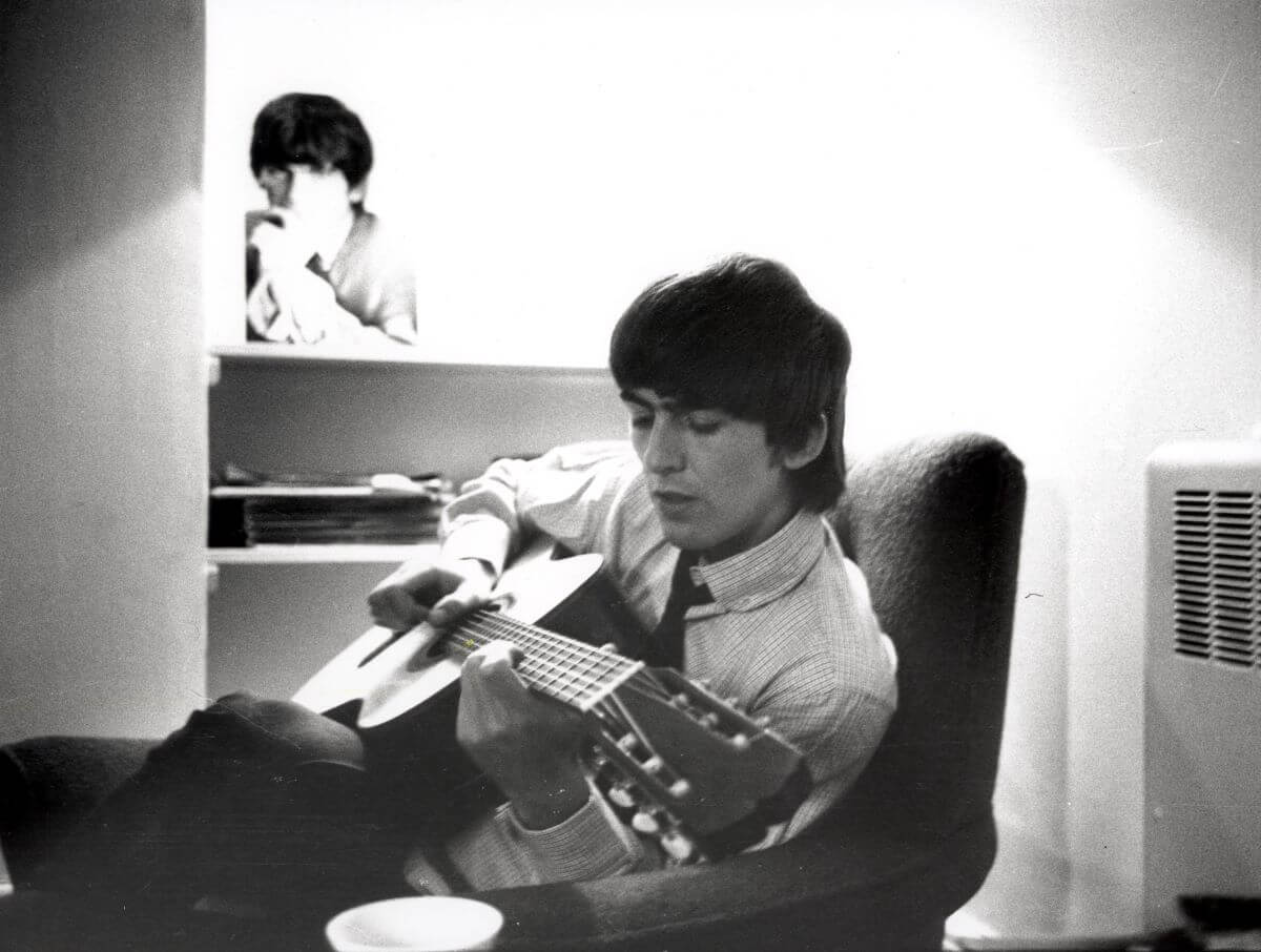 A black and white picture of The Beatles' George Harrison sitting in a chair with an acoustic guitar.
