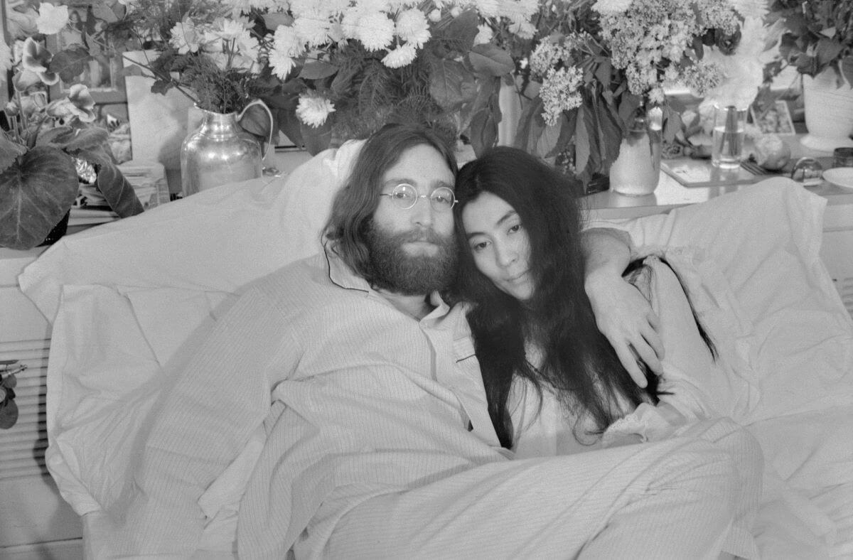 A black and white picture of John Lennon and Yoko Ono in bed together. He has his arm around her shoulders.