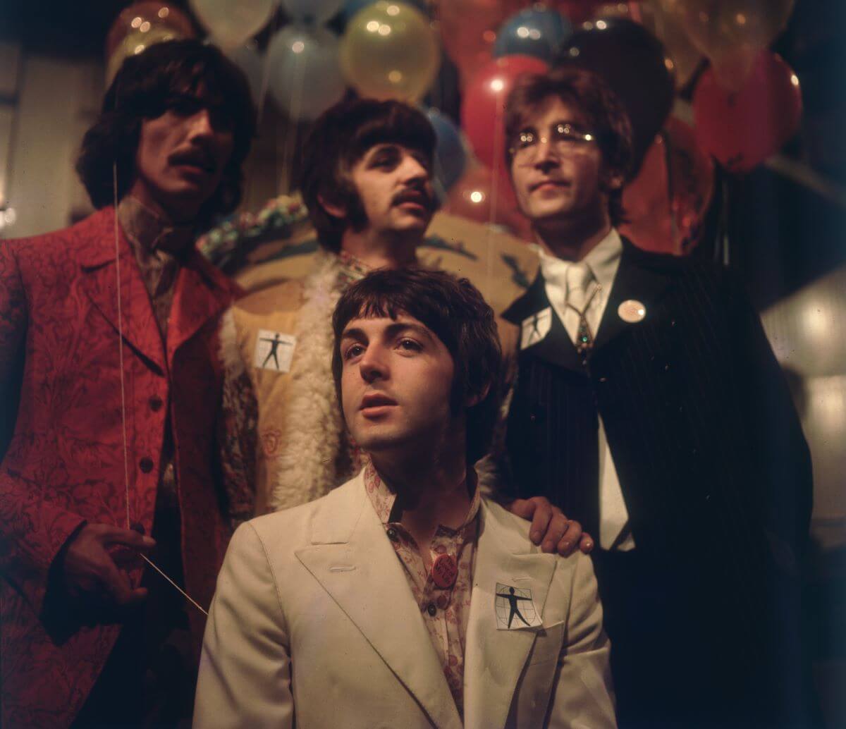 George Harrison, Ringo Starr, and John Lennon stand behind Paul McCartney, who sits. There are balloons behind them.