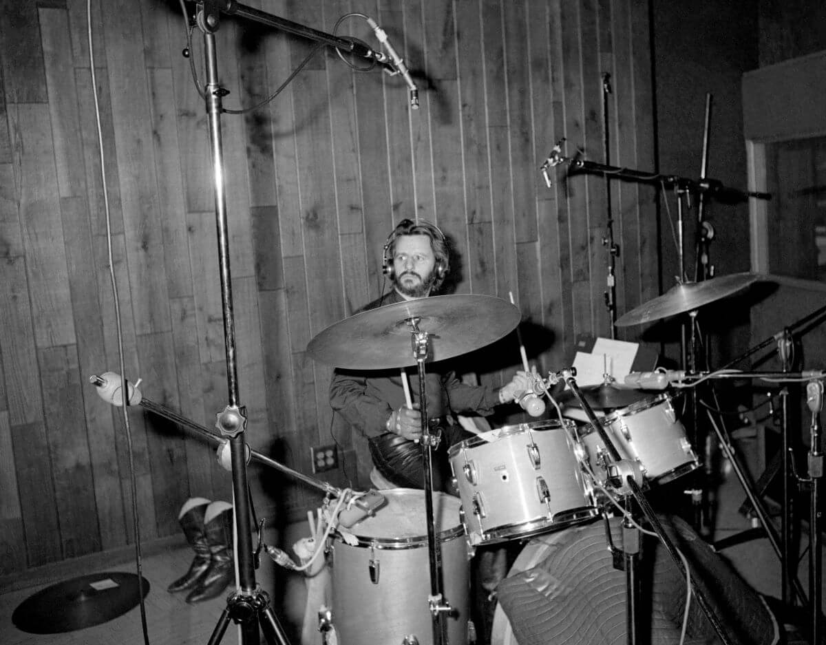 A black and white picture of Ringo Starr sitting at a drum set in front of a wood paneled wall.