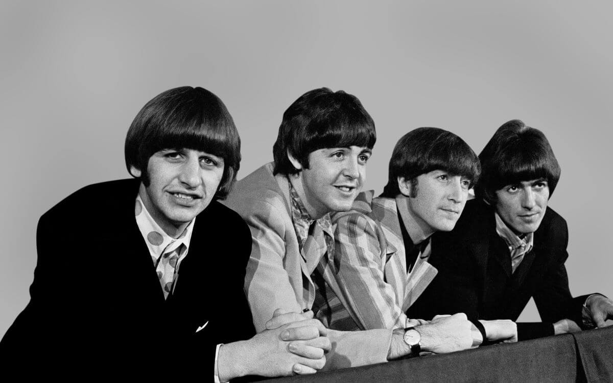 A black and white picture of Ringo Starr, Paul McCartney, John Lennon, and George Harrison of The Beatles. They pose in a line and Starr looks into the camera.