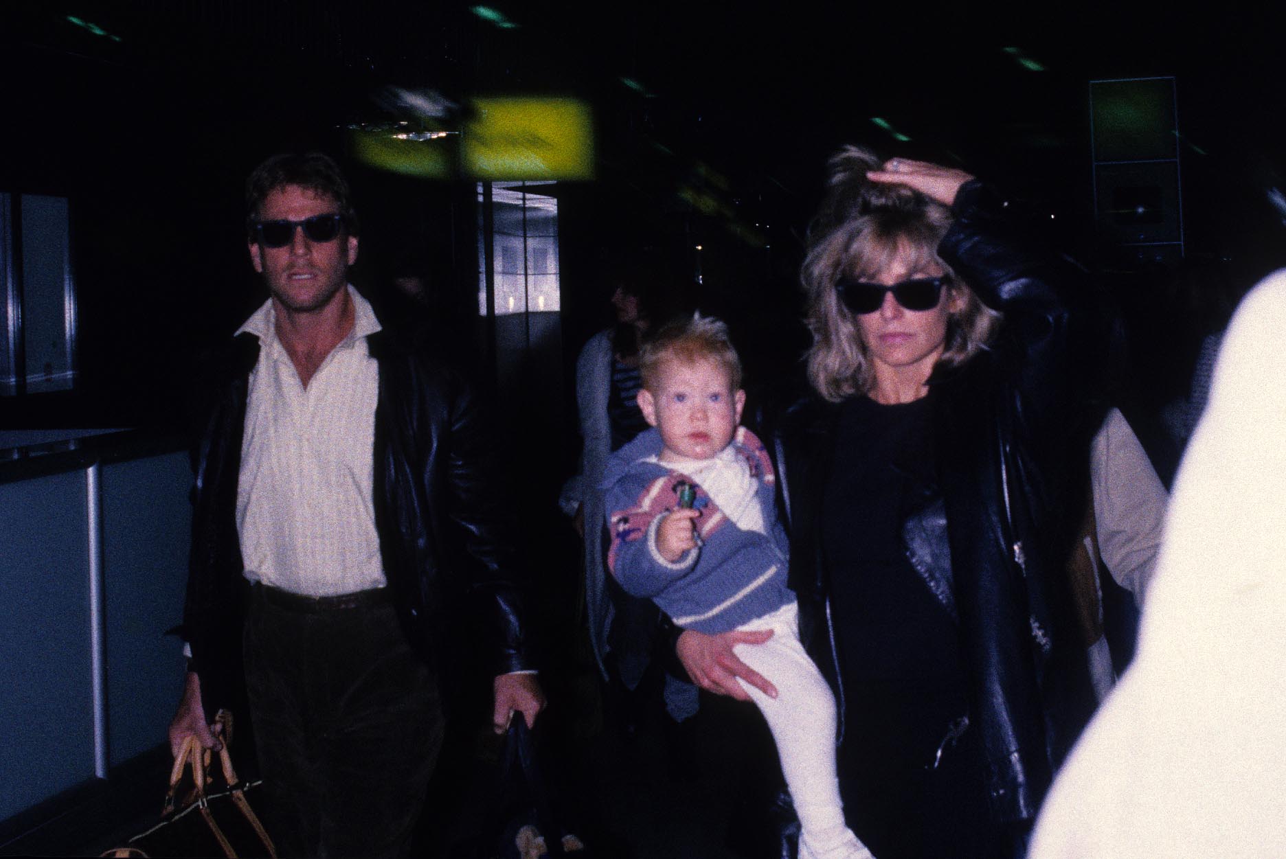 Ryan O'Neal in sunglasses walking next to Farrah Fawcett, who is holding their son Redmond