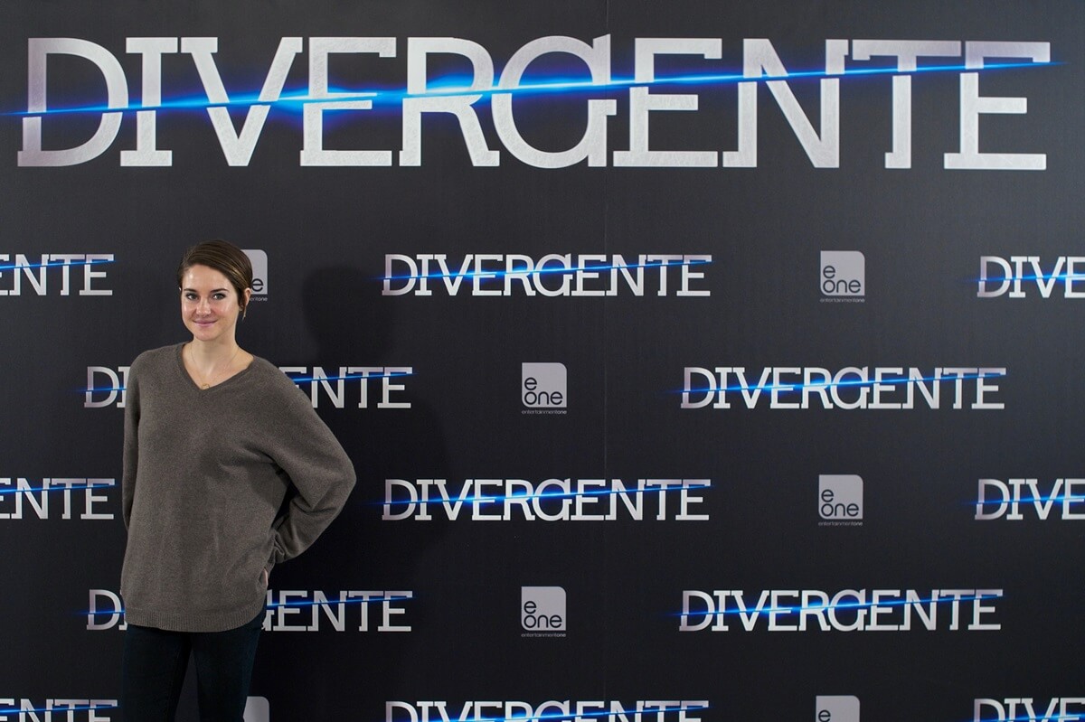 Shailene Woodley posing in front of a Divergent poster while wearing grey and black clothes.