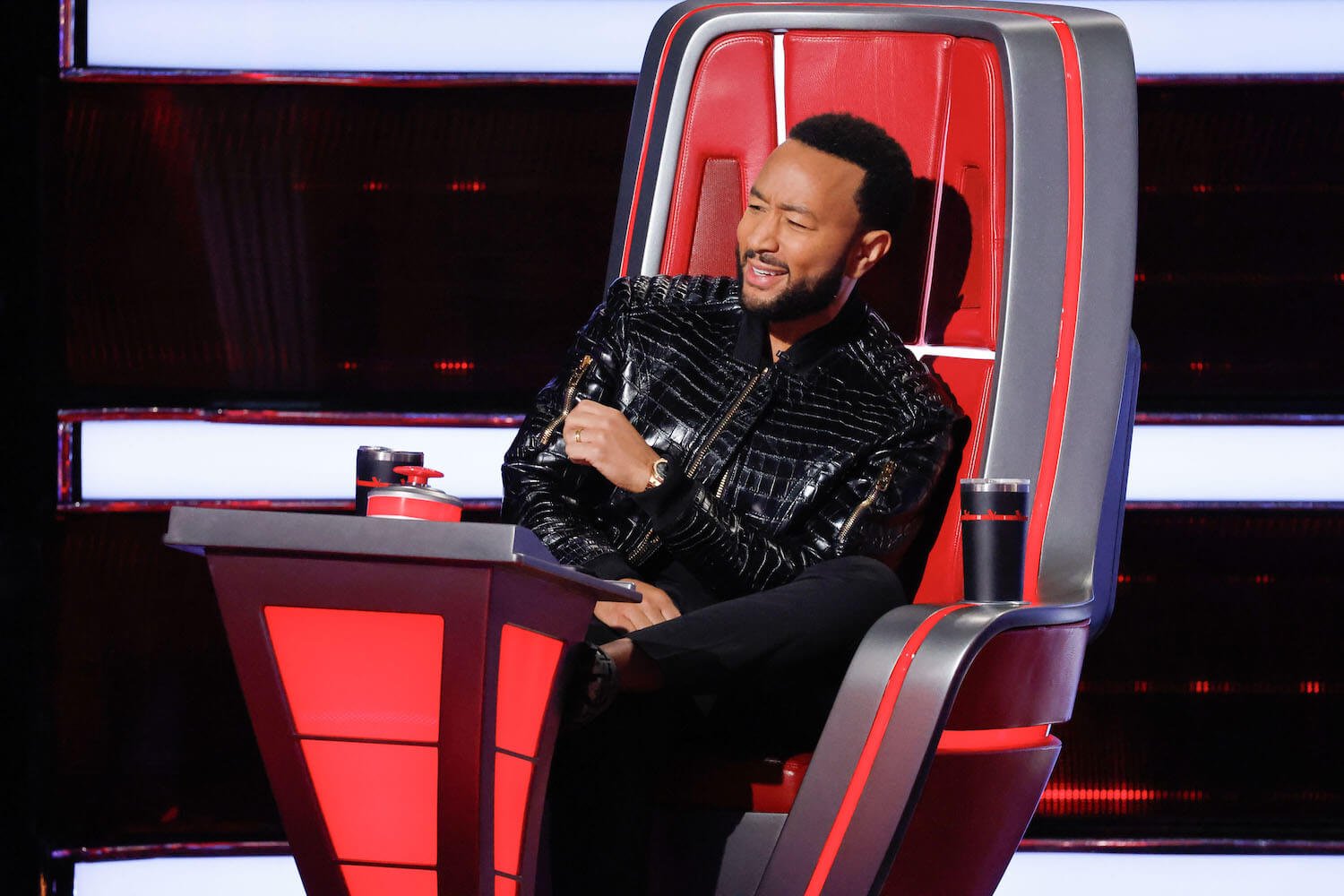 John Legend sitting in a coaches chair laughing in 'The Voice' Season 24