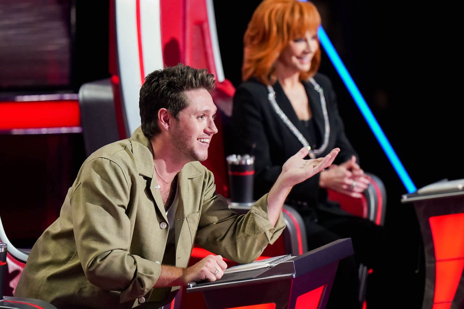 'The Voice' Season 24 coach Niall Horan sitting in a coach chair gesturing with Reba McEntire blurry behind him