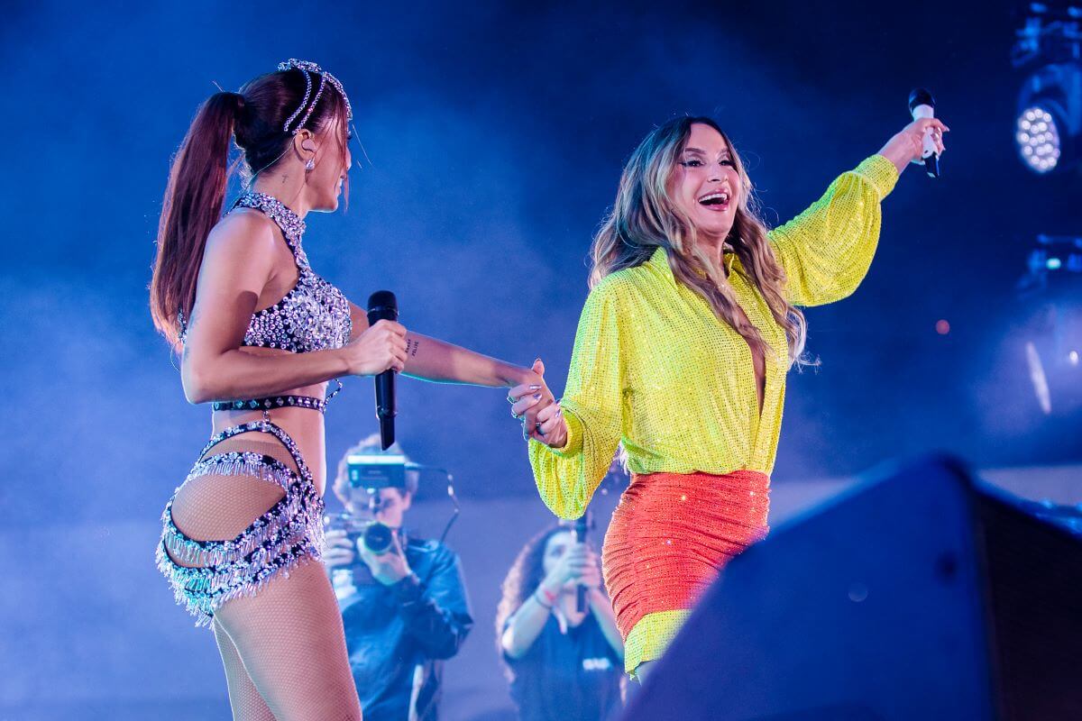 Anitta holds a microphone and stands next to Claudia Leitte onstage. Anitta wears a silver bodysuit and Leitte wears a neon yellow shirt and red skirt.