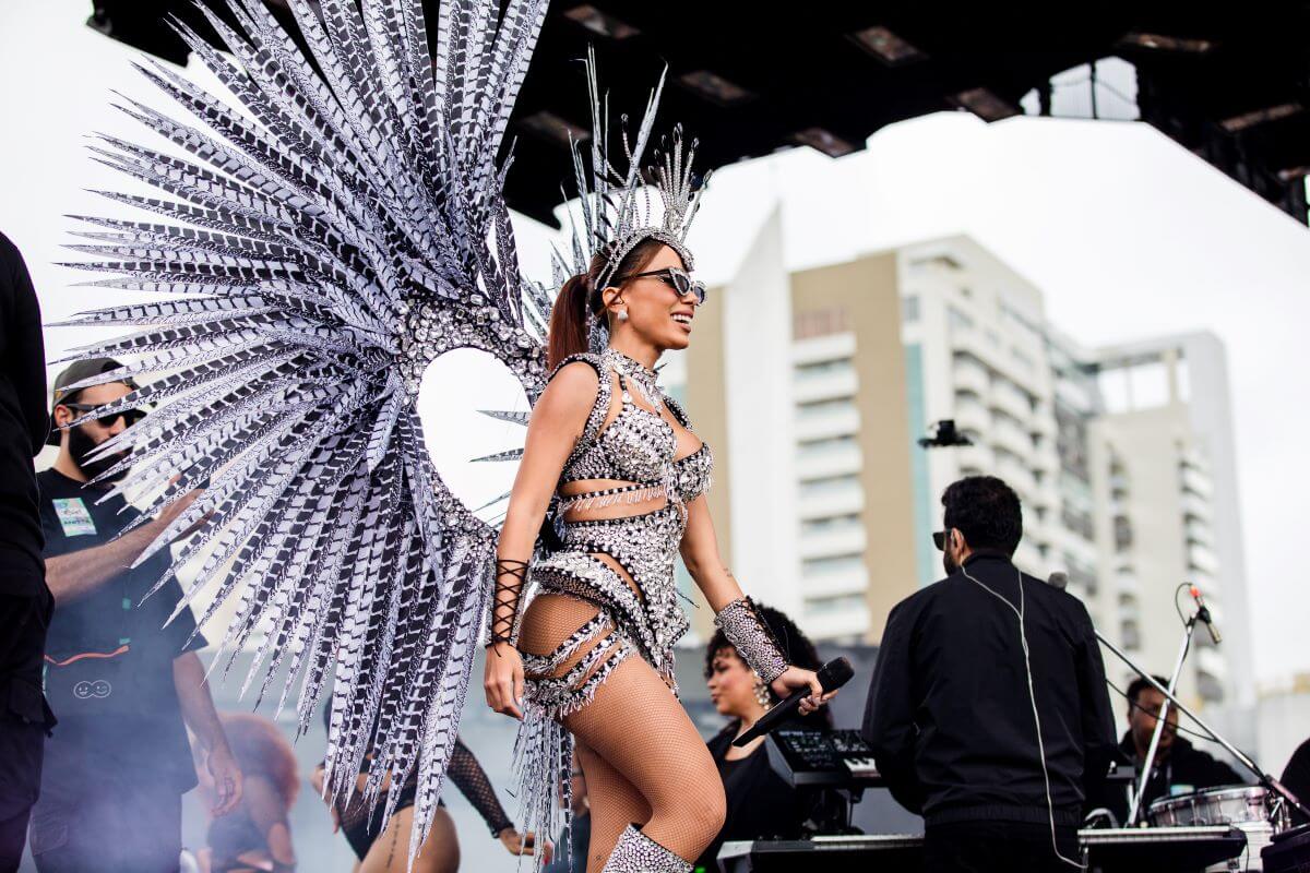 Anita wears a silver bodysuit with matching wings, boots, sunglasses, and a headdress. She stands on a stage.