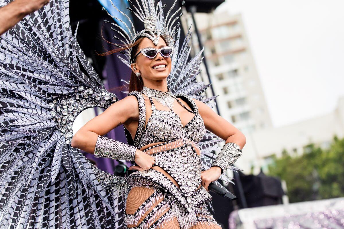 Anitta wears a bodysuit, sunglasses, and wings and places her hands on her hips.