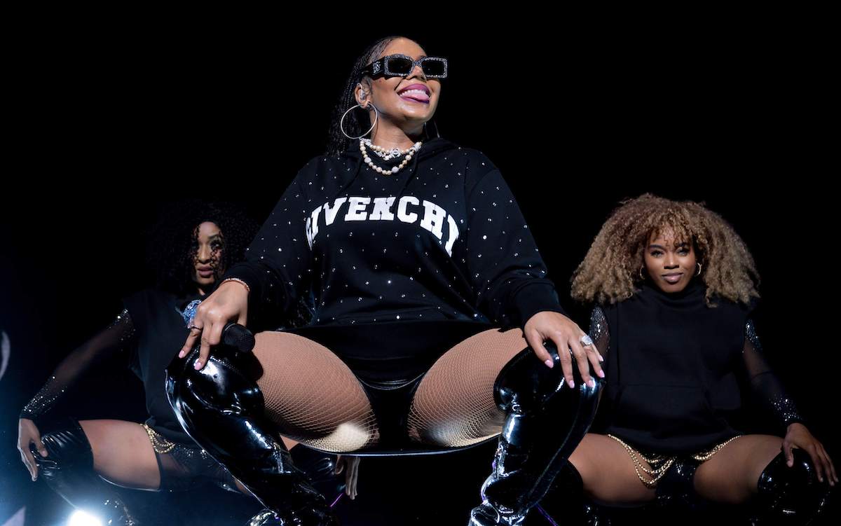 Singer Ashanti squats on stage in a black sweatshirt while her backup dancers squat behind her