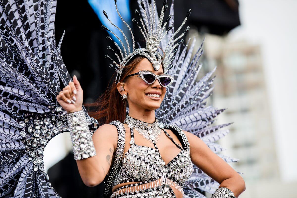 Brazilian pop star Anitta wears a silver jumpsuit, wings, headdress, and sunglasses. She holds one hand up and has the other on her hip.