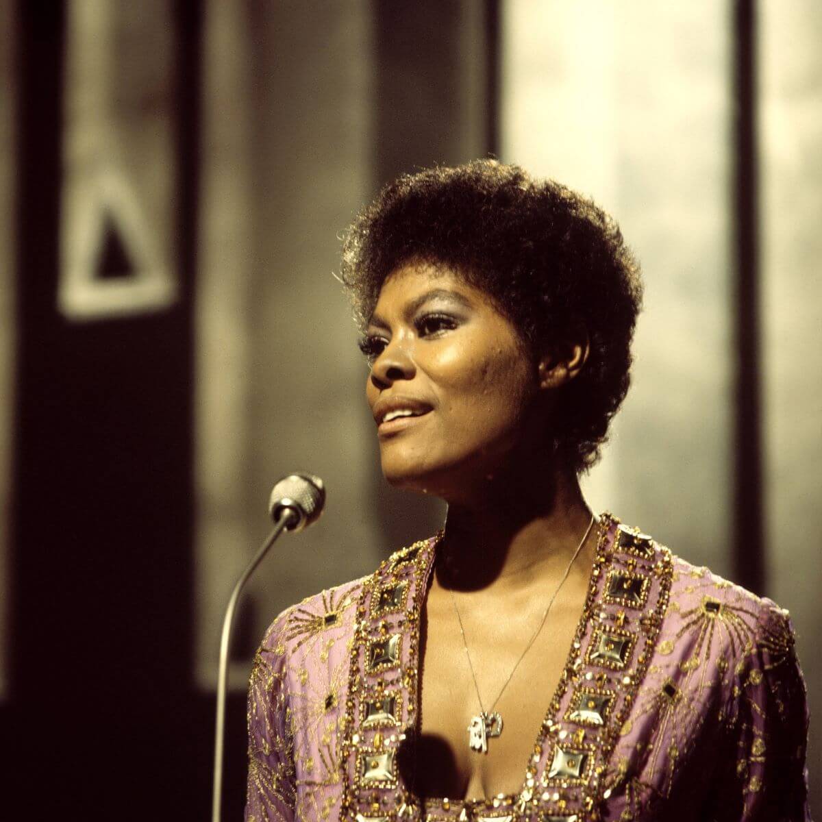 Dionne Warwick wears a purple jeweled dress and stands in front of a microphone.
