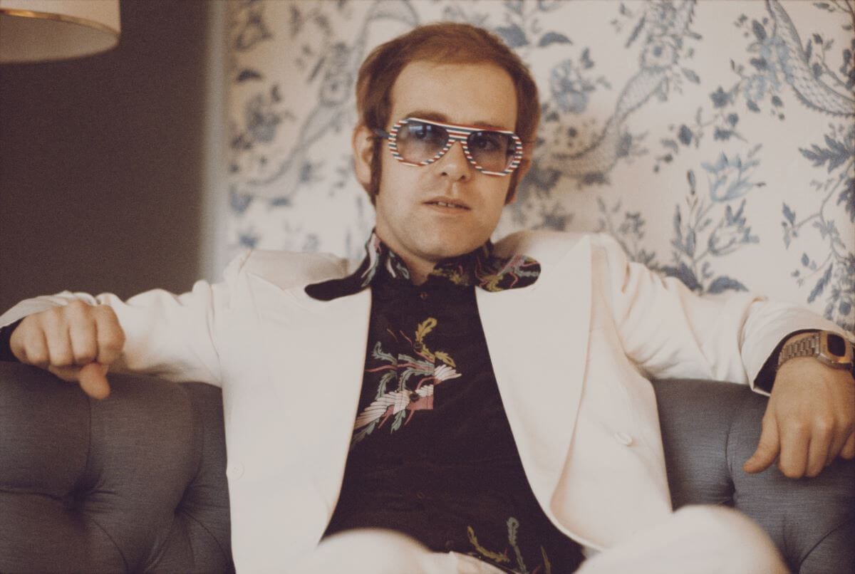 Elton John wears sunglasses and sits in an armchair.
