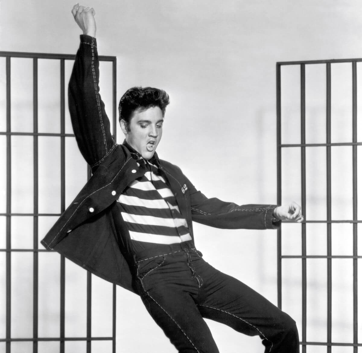 A black and white picture of Elvis wearing a striped shirt and dancing. He lifts one arm above his head.