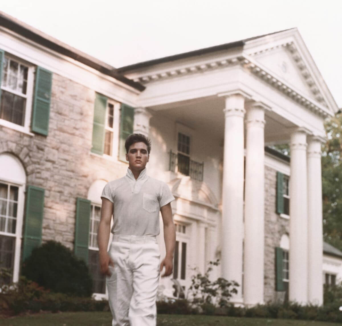 Elvis wears white pants and a white shirt and walks in front of Graceland.