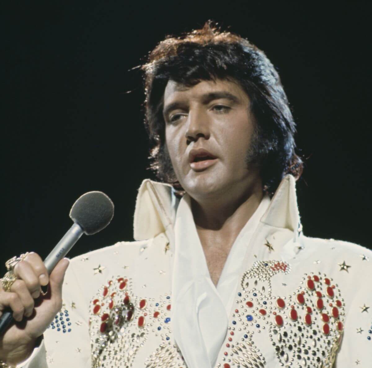 Elvis Presley wears a bedazzled white jumpsuit and holds a microphone.