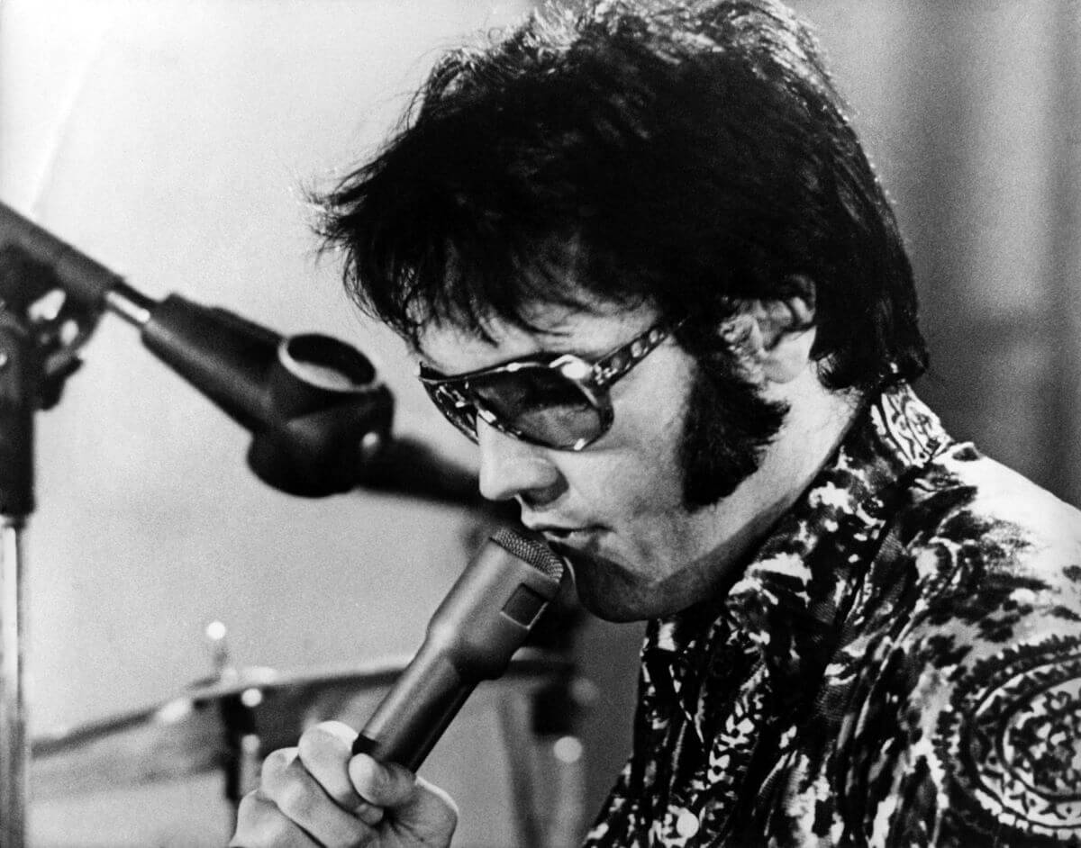 A black and white picture of Elvis wearing sunglasses and holding a microphone to his mouth.