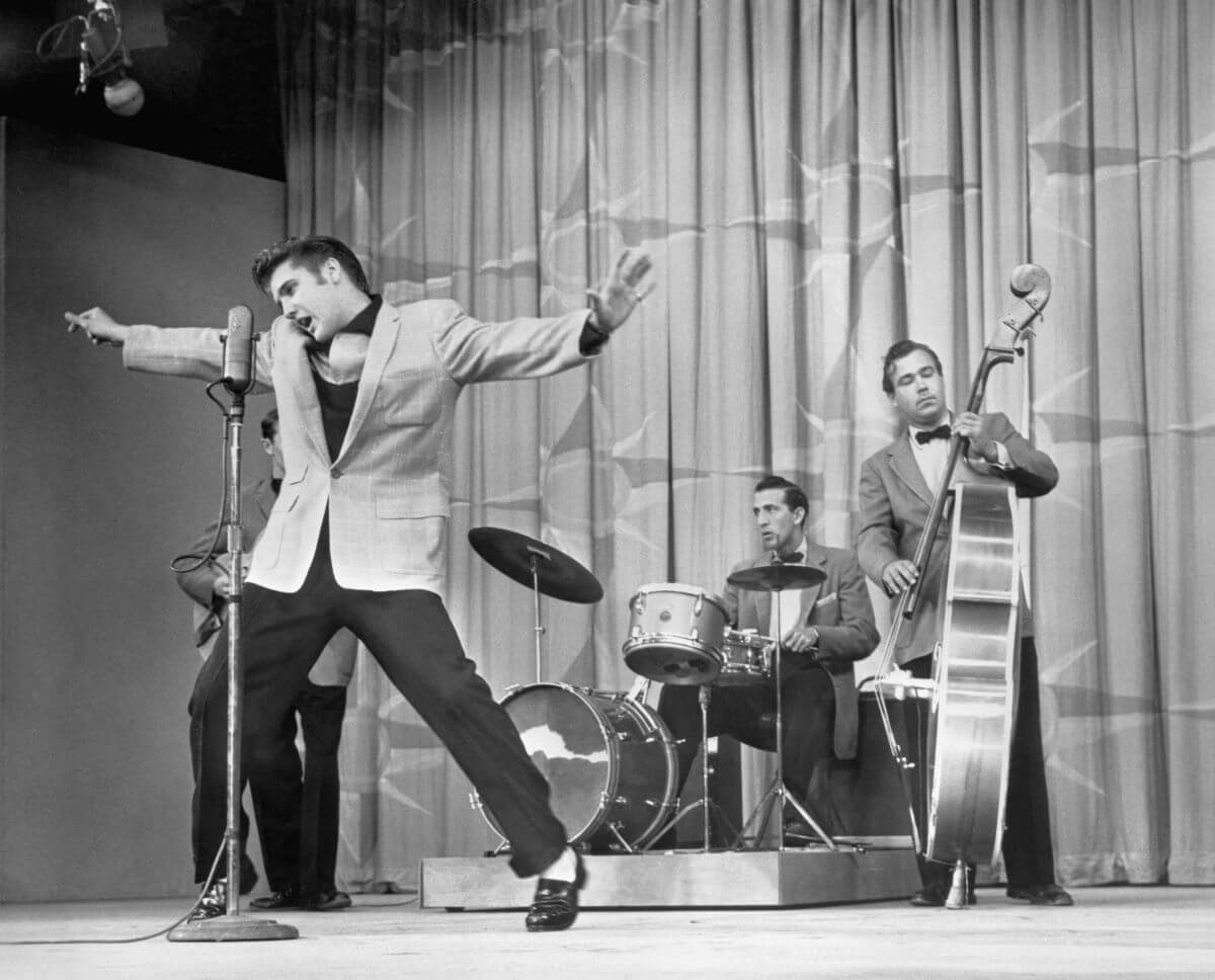 A black and white picture of Elvis standing in front of a microphone with his arms spread wide. A drummer and a bass player stand behind him.