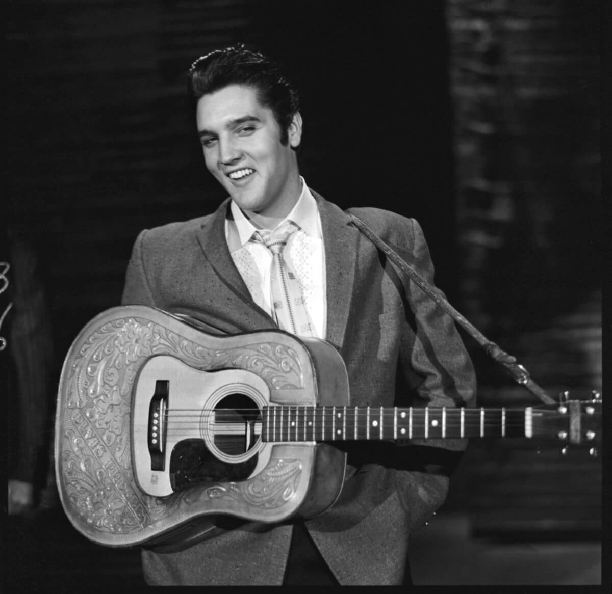 A black and white picture of Elvis wearing a suit and holding an acoustic guitar. He smiles.