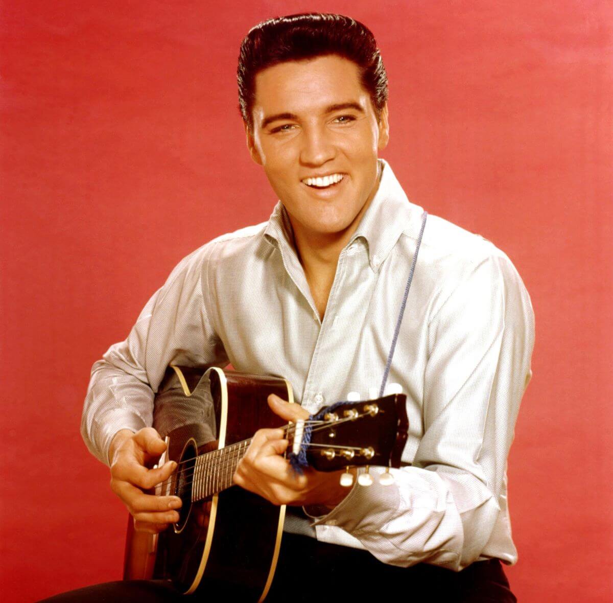 Elvis Presley wears a white shirt and sits with an acoustic guitar. He is in front of a red background.