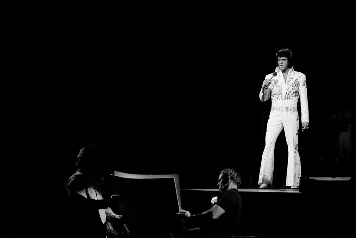 A black and white picture of Elvis Presley wearing a white jumpsuit and standing onstage.