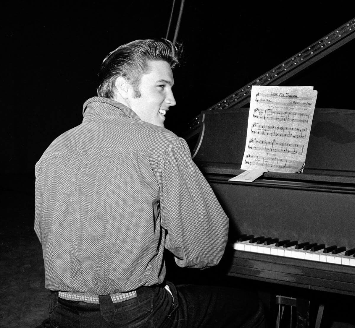 A black and white picture of Elvis Presley sitting at a piano with his back to the camera.