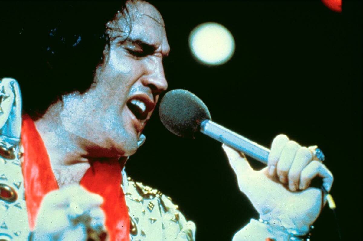 Elvis wears a white jumpsuit and red scarf. He sings into a microphone.