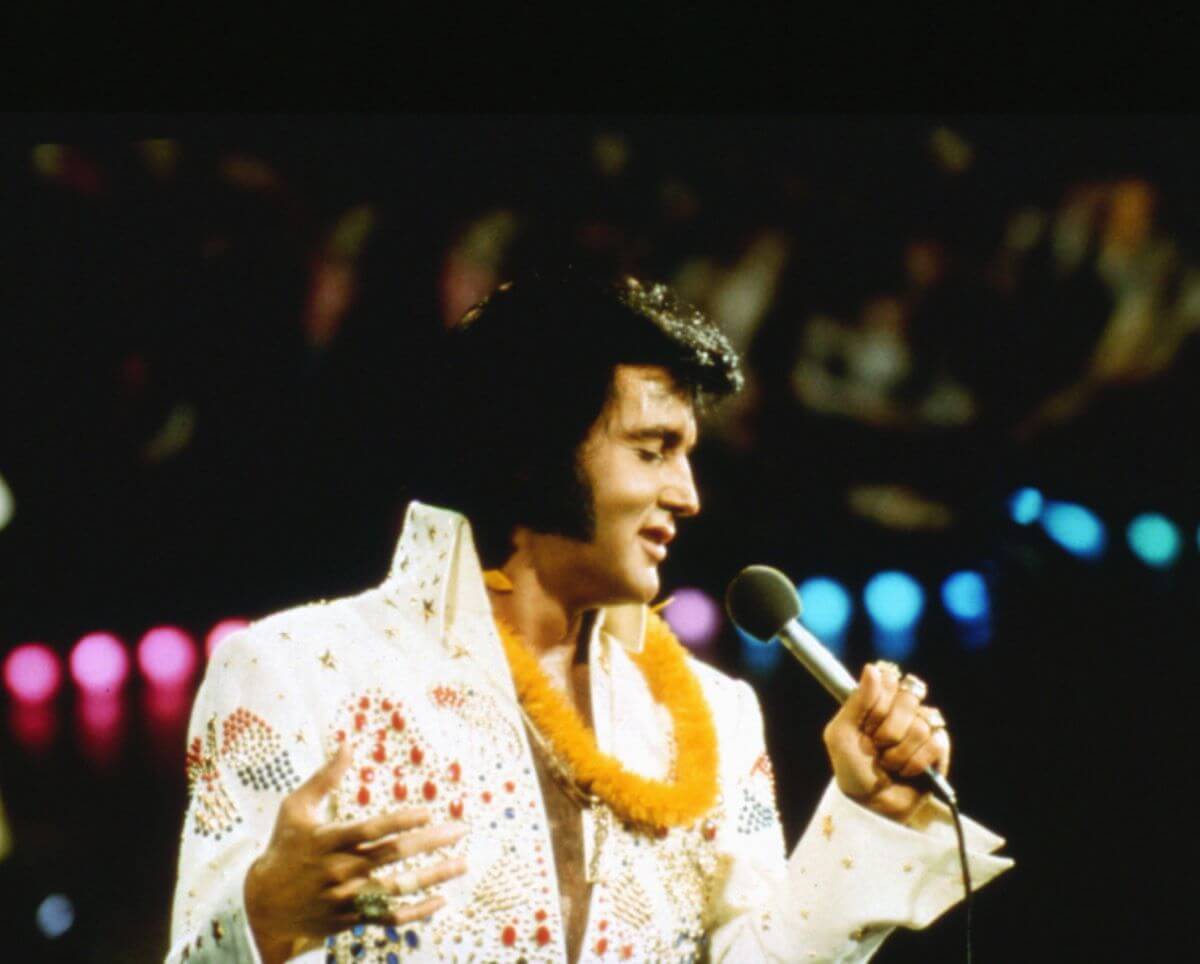 Elvis wears a white jumpsuit with a yellow lei around his neck. He sings into a microphone.