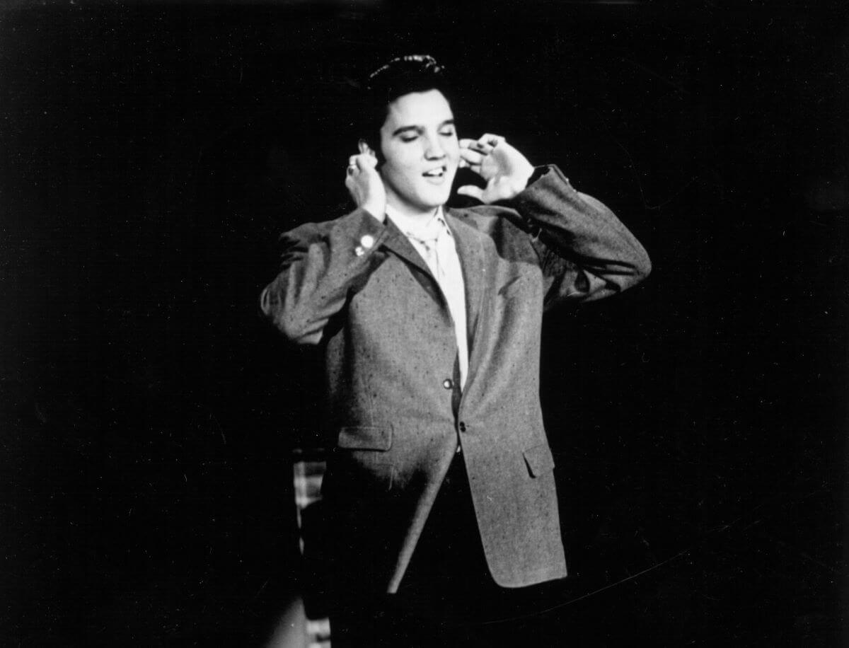 A black and white picture of Elvis Presley lifting his hands to his ears while he sings.