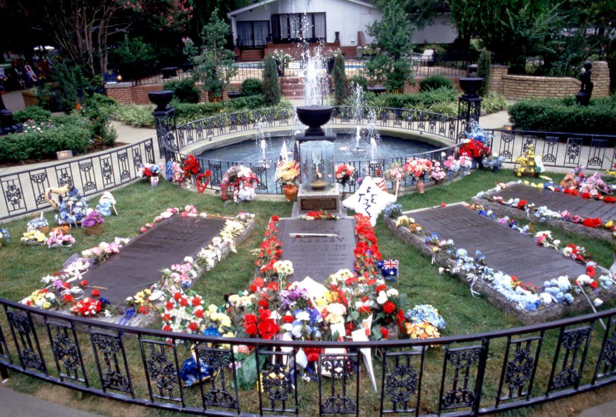Four headstones at Graceland lie with flowers around them. There is a fountain and a pool house behind them.